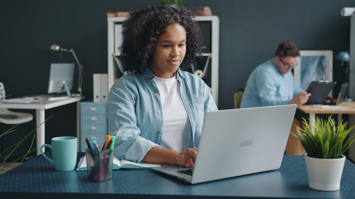 A young professional working on a laptop at a tidy desk in a modern open-plan office, a colleague in the background.