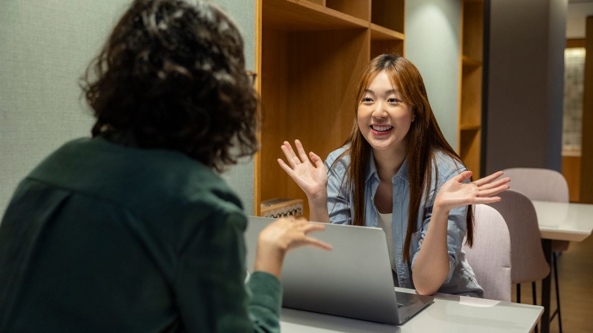 A manager and an employee having an animated one-on-one conversation across a desk with a laptop between them.
