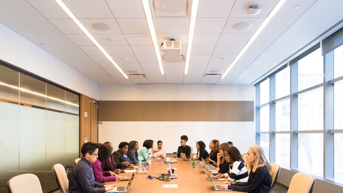 A group of colleagues seated around a long conference table with laptops in a bright meeting room.